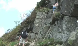 Turysta z plecakiem wspina się po skalistej ścieżce, obok wspina się inna osoba.