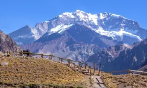 Majestatyczne góry z ośnieżonym szczytem Aconcagua w tle. Ścieżka z drewnianym ogrodzeniem prowadzi przez suchą, trawiastą górę.