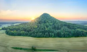Wschód słońca nad zielonym wzgórzem, przypominającym kształtem wygasły wulkan. W oddali rozciąga się krajobraz pól i lasów.