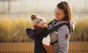 Uśmiechnięta mama z dzieckiem w nosidełku dla dziecka w góry. Ciepłe ubrania i zimowa czapka chronią przed chłodem.