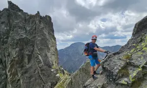 Wspinacz na via ferracie, z plecakiem i kaskiem, trzyma się stalowej liny. W tle skaliste szczyty i chmury. Brak czerwonej ławki.