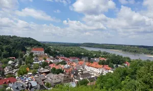Widok na Kazimierz Dolny z górami, rzeką Wisłą i charakterystyczną zabudową. W oddali widać wzgórza, a na pierwszym planie urokliwe kamienice.