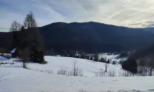 Zimowy krajobraz z ośnieżonymi polami i lasem u podnóża masywnej, ciemnej góry. W oddali widać kilka domów. Jasień góra wita zimowym spokojem.