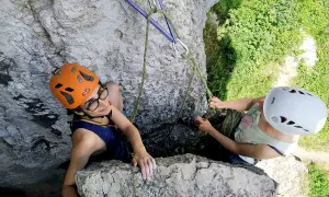 Dwie dziewczyny w kaskach podczas wspinaczki skalnej. Jedna z nich, w pomarańczowym kasku, uśmiecha się do obiektywu, druga, w białym kasku, asekuruje ją.