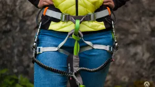 Osoba w kamizelce i jeansach ma na sobie uprząż z lonżą via ferrata, gotowa do wspinaczki.