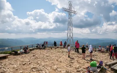 Ludzie odpoczywają na szczycie, podziwiając panoramę Bieszczad. Metalowy krzyż góruje nad kamienistym terenem, świadcząc o popularności tego miejsca na szlaki Bieszczady.