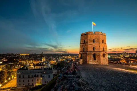 Wieża na wzgórzu Giedymina z flagą Litwy na tle zachodzącego słońca i oświetlonego Wilna.