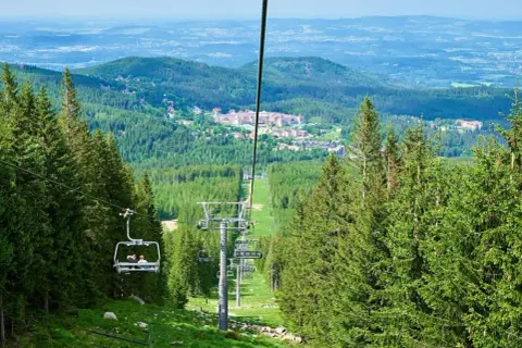 Wjazd kolejką na wysoką kopę, otoczoną lasem świerkowym. W oddali widać zabudowania i górzysty krajobraz.