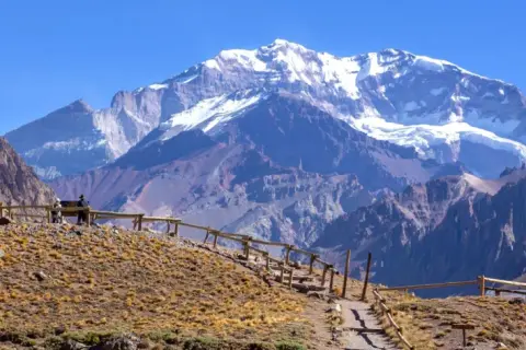 Majestatyczne góry z ośnieżonym szczytem Aconcagua w tle. Ścieżka z drewnianym ogrodzeniem prowadzi przez suchą, trawiastą górę.