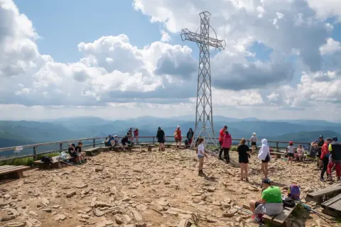 Ludzie odpoczywają na szczycie, podziwiając panoramę Bieszczad. Metalowy krzyż góruje nad kamienistym terenem, świadcząc o popularności tego miejsca na szlaki Bieszczady.