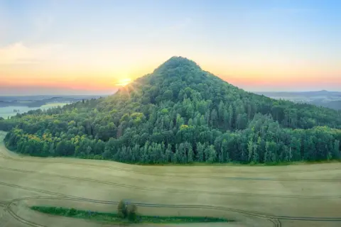 Wschód słońca nad zielonym wzgórzem, przypominającym kształtem wygasły wulkan. W oddali rozciąga się krajobraz pól i lasów.