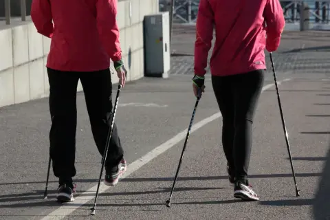 Dwie osoby w czerwonych kurtkach i czarnych spodniach uprawiają nordic walking na asfaltowej ścieżce.