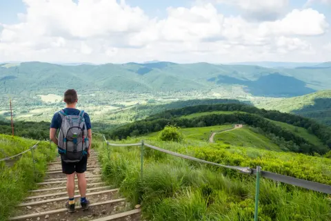 Mężczyzna z plecakiem schodzi po kamiennych schodach. Przed nim rozciąga się malowniczy szlak na Tarnicę, otoczony zielonymi wzgórzami i błękitnym niebem.
