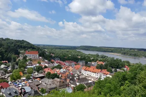 Widok na Kazimierz Dolny z górami, rzeką Wisłą i charakterystyczną zabudową. W oddali widać wzgórza, a na pierwszym planie urokliwe kamienice.