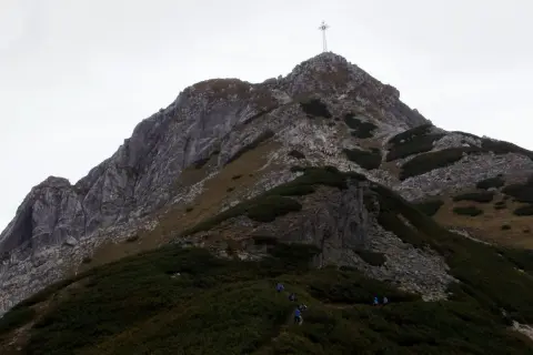 Grupa turystów wspina się po skalistej ścieżce na Giewont. Widoczny krzyż na szczycie. Trasa jest wymagająca.