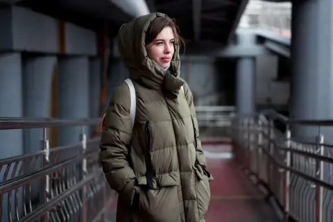 Kobieta w zielonej, pikowanej parkce z kapturem, stojąca na tle miejskiej architektury. Parka co to? To idealna ochrona przed zimnem.