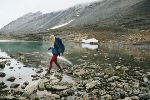 Kobieta z plecakiem przemierza kamieniste wybrzeże jeziora. W tle góry, idealne dla skandynawskie marki outdoorowe.