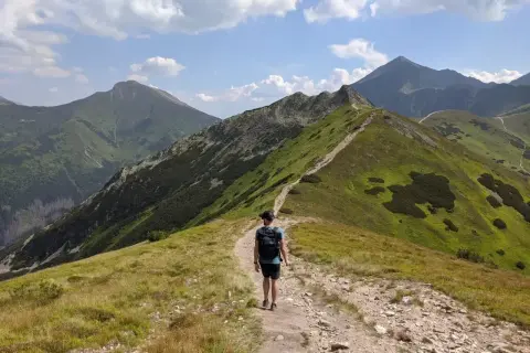 Wędrowiec na szlaku prowadzącym na **ornak szczyt**. Zielone zbocza górskie, kamienista ścieżka i błękitne niebo z chmurami.