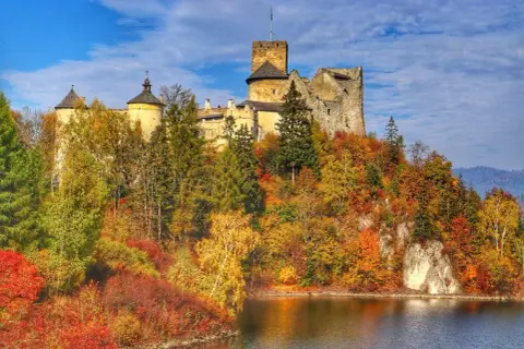 Zamek Dunajec w jesiennej szacie, otoczony kolorowymi drzewami, góruje nad spokojną taflą wody.