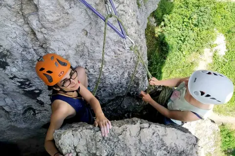 Dwie dziewczyny w kaskach podczas wspinaczki skalnej. Jedna z nich, w pomarańczowym kasku, uśmiecha się do obiektywu, druga, w białym kasku, asekuruje ją.