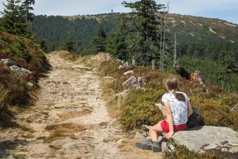 Dziewczynka odpoczywa na kamieniu przy kamienistej ścieżce. W oddali widać las i wieżę widokową. To piękna **śnieżnik trasa**.