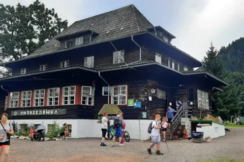 Drewniana chata "Andrzejówka" u podnóża **waligóra szczyt**. Ludzie odpoczywają przed wejściem, a w tle widać gęsty las.