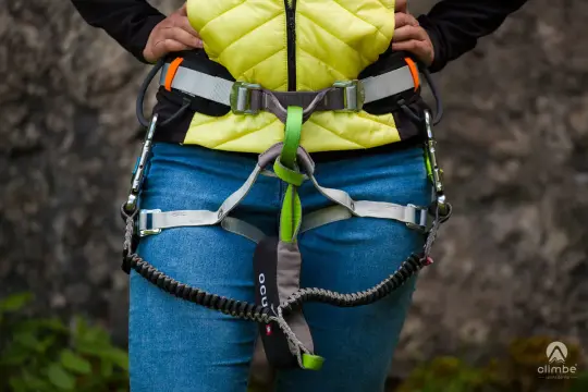 Osoba w kamizelce i jeansach ma na sobie uprząż z lonżą via ferrata, gotowa do wspinaczki.