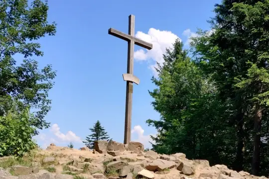 Drewniany krzyż na skalistym szczycie, punkt orientacyjny na łysica szlak. Otoczony zielenią drzew i błękitem nieba z chmurami.