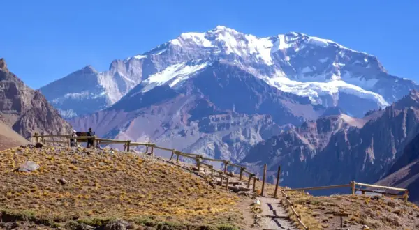 Majestatyczne góry z ośnieżonym szczytem Aconcagua w tle. Ścieżka z drewnianym ogrodzeniem prowadzi przez suchą, trawiastą górę.