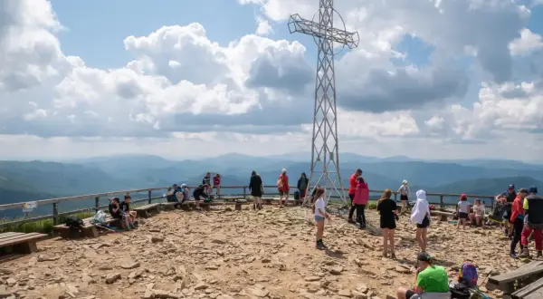 Ludzie odpoczywają na szczycie, podziwiając panoramę Bieszczad. Metalowy krzyż góruje nad kamienistym terenem, świadcząc o popularności tego miejsca na szlaki Bieszczady.