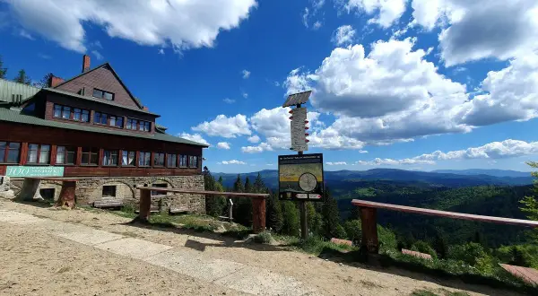 Drewniane schronisko na szczycie, z którego roztacza się widok na gęsto zalesione góry. Obok drogowskaz i ławki. Stożek wielki w oddali.