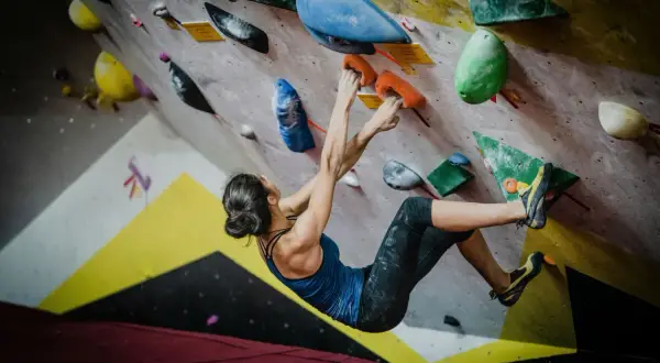 Kobieta wspina się po kolorowej ścianie, chwytając się pomarańczowych uchwytów. To właśnie bouldering – sportowa wspinaczka bez lin.