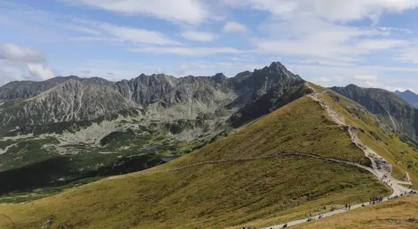 Tłum turystów wędruje kamienistym szlakiem na Kasprowy Wierch. Widok na skaliste szczyty Tatr i zielone doliny.
