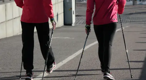 Dwie osoby w czerwonych kurtkach i czarnych spodniach uprawiają nordic walking na asfaltowej ścieżce.