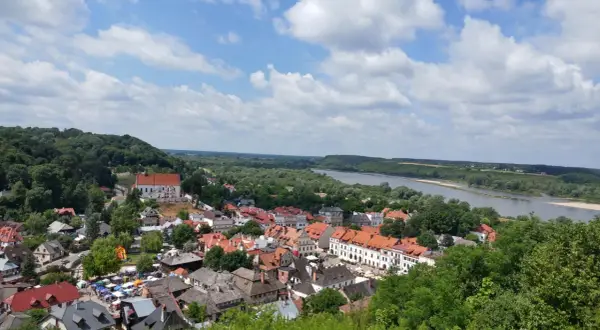 Widok na Kazimierz Dolny z górami, rzeką Wisłą i charakterystyczną zabudową. W oddali widać wzgórza, a na pierwszym planie urokliwe kamienice.