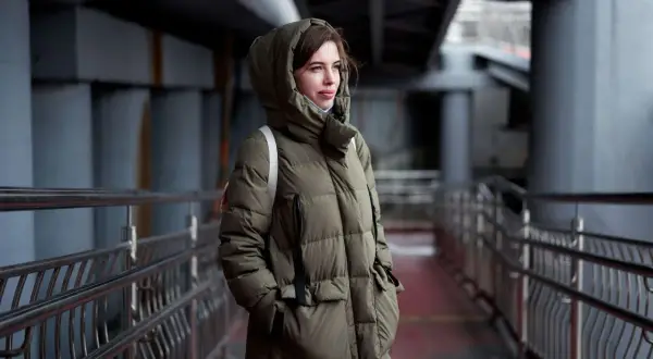 Kobieta w zielonej, pikowanej parkce z kapturem, stojąca na tle miejskiej architektury. Parka co to? To idealna ochrona przed zimnem.