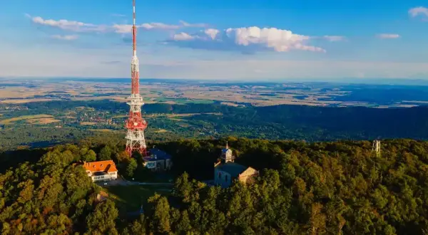 Wieża RTV na górze Ślęża góruje nad lasami i krajobrazem. Obok niej budynki i kościółek.
