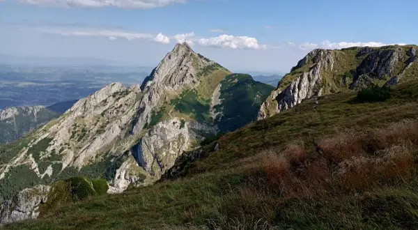 Widok na skaliste szczyty i zielone zbocza. Na pierwszym planie trawy, w oddali dolina małej łąki.