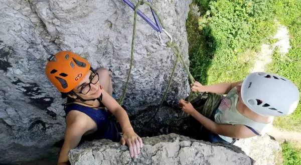 Dwie dziewczyny w kaskach podczas wspinaczki skalnej. Jedna z nich, w pomarańczowym kasku, uśmiecha się do obiektywu, druga, w białym kasku, asekuruje ją.