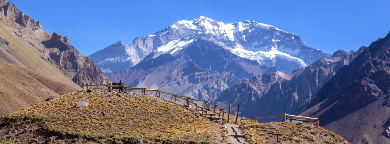 Majestatyczne góry z ośnieżonym szczytem Aconcagua w tle. Ścieżka z drewnianym ogrodzeniem prowadzi przez suchą, trawiastą górę.