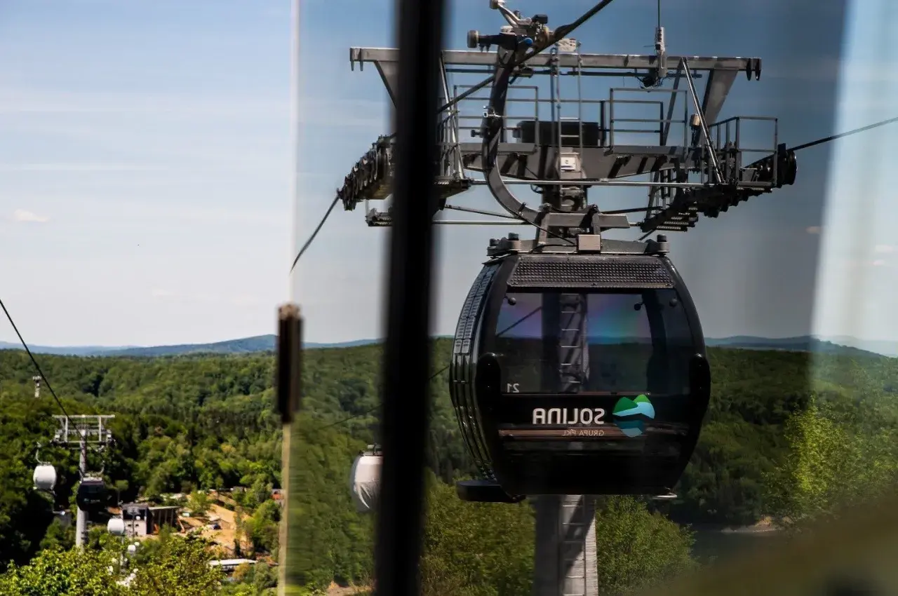 Widok z okna na solina kolejka linowa. Czarna gondola z logo "Solina" wznosi się nad zielonymi lasami i wzgórzami.