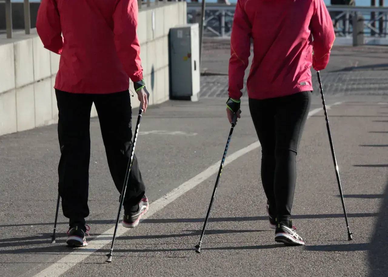 Dwie osoby w czerwonych kurtkach i czarnych spodniach uprawiają nordic walking na asfaltowej ścieżce.