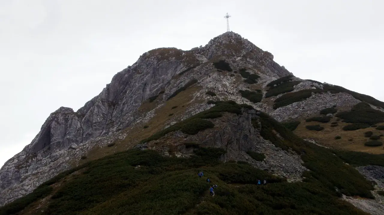 Grupa turystów wspina się po skalistej ścieżce na Giewont. Widoczny krzyż na szczycie. Trasa jest wymagająca.