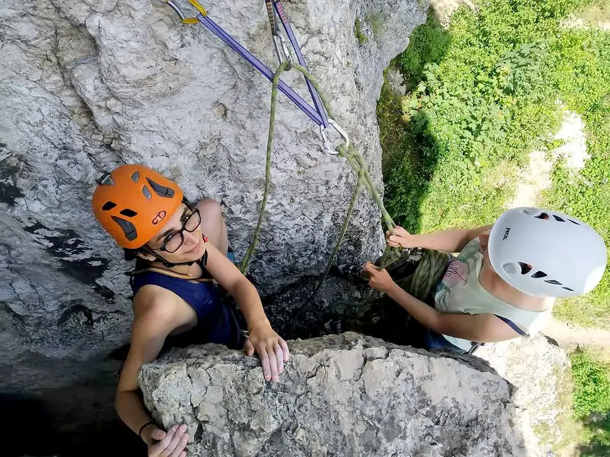 Dwie dziewczyny w kaskach podczas wspinaczki skalnej. Jedna z nich, w pomarańczowym kasku, uśmiecha się do obiektywu, druga, w białym kasku, asekuruje ją.