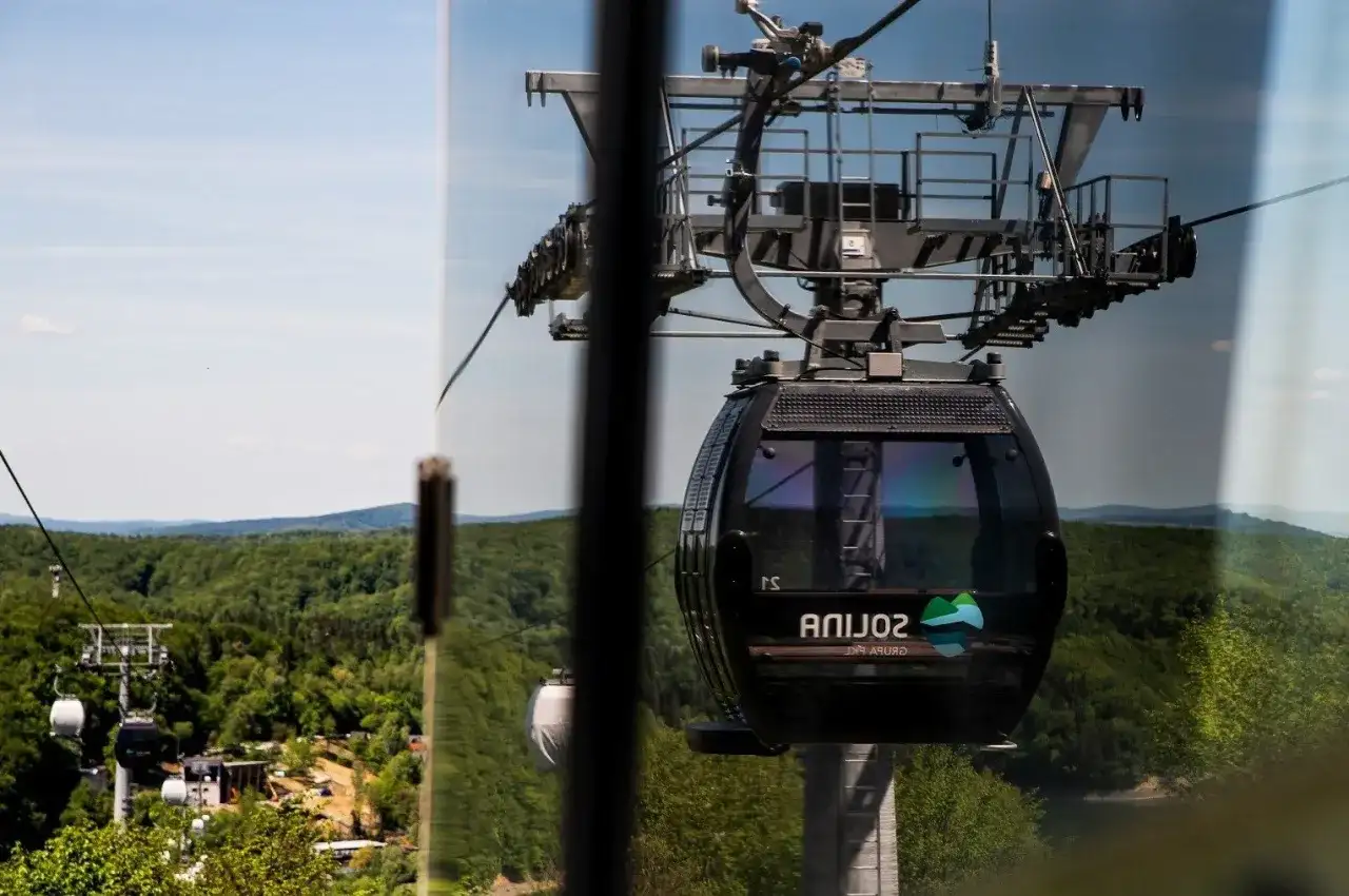 Widok z okna na solina kolejka linowa. Czarna gondola z logo "Solina" wznosi się nad zielonymi lasami i wzgórzami.