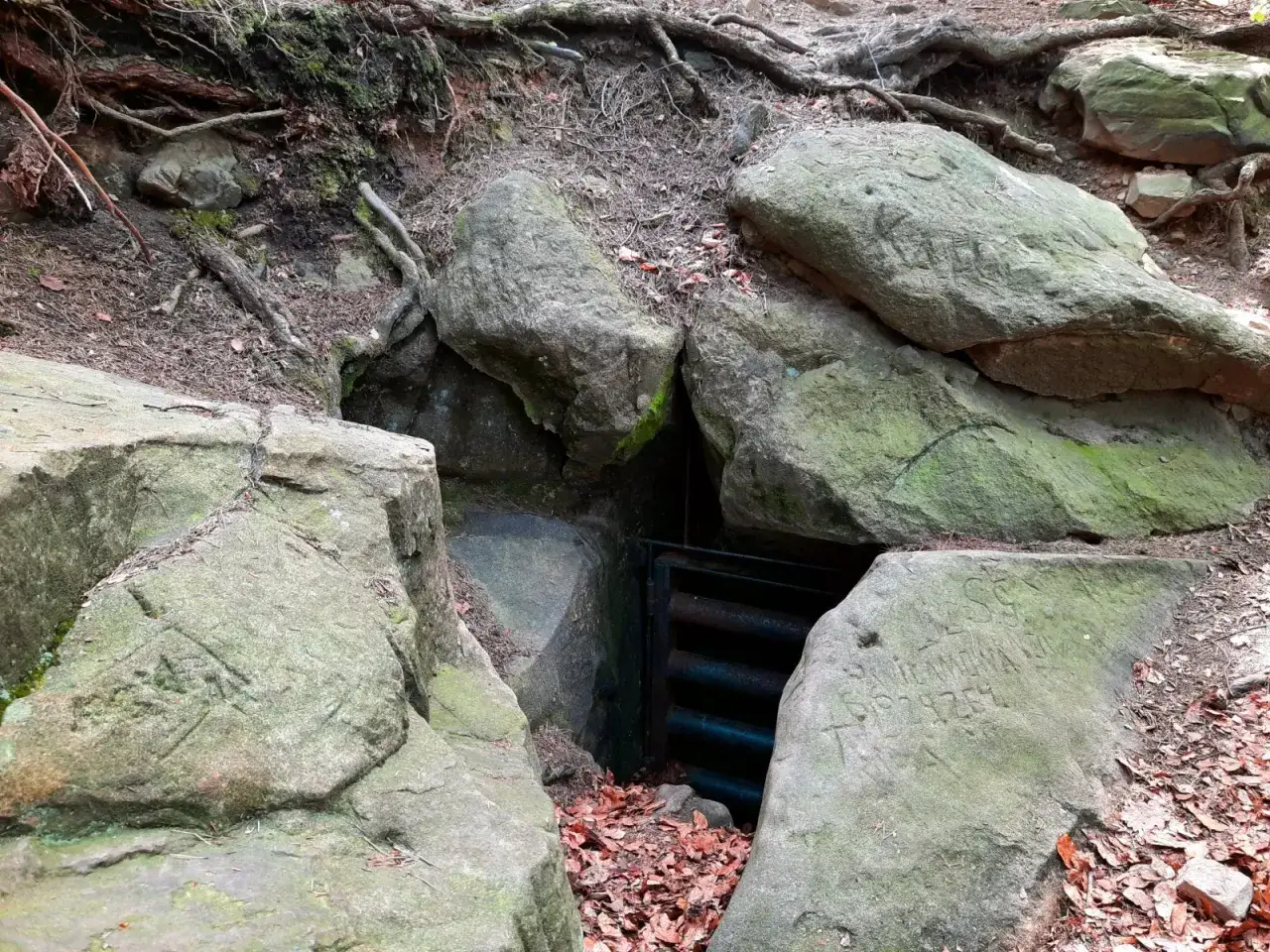 Wąskie przejście między skalnymi blokami, zwanymi diable skały, prowadzi do ciemnego wnętrza z metalową kratą.