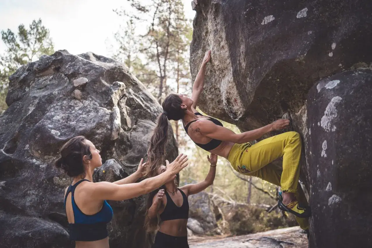 Trzy kobiety uprawiają bouldering. Jedna wspina się po skale, dwie inne asekurują ją.