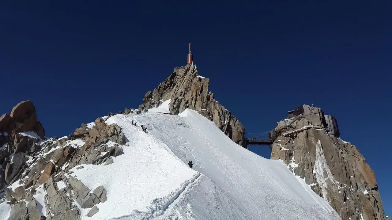 Wspinacze na śnieżnym zboczu Mont Blanc, gdzie leży ten majestatyczny szczyt Alp. W oddali widać budynki i wieżę.