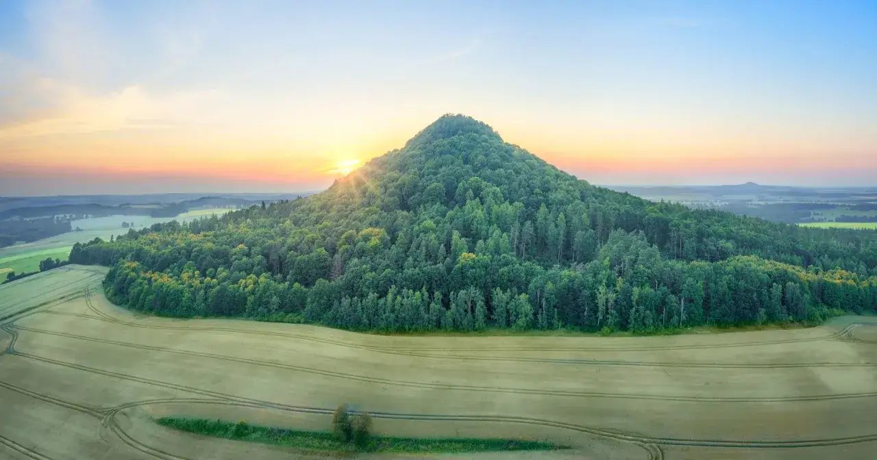 Wsch&oacute;d słońca nad zielonym wzg&oacute;rzem, przypominającym kształtem wygasły wulkan. W oddali rozciąga się kraina wygasłych wulkan&oacute;w, porośnięta lasami i polami.