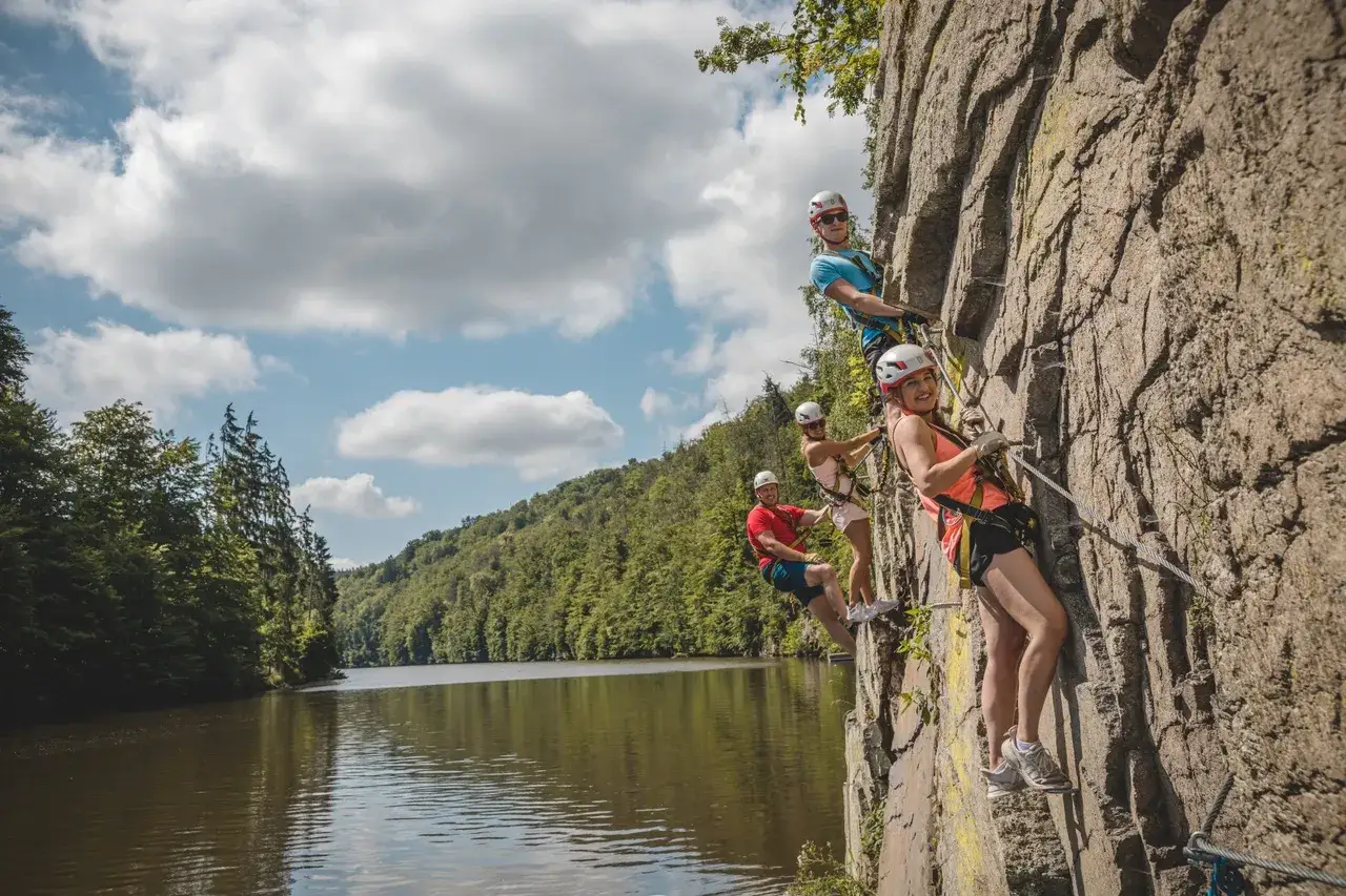 Grupa ludzi wspina się po skale na via ferrata w Czechach. W tle jezioro i zielone wzg&oacute;rza.